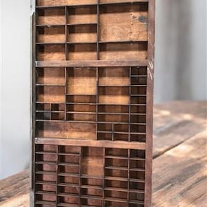 May include: A vintage wooden printer's tray with numerous small compartments of varying sizes. The tray has a rectangular shape with a dark wood finish and a metal frame. The compartments are designed to hold individual letters or other small items.
