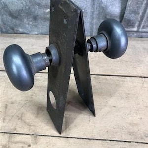 Könnte beinhalten: Ein Vintage-Türknopf-Set mit zwei runden Knöpfen und einer Metall-Rückplatte. Die Knöpfe sind dunkelgrau und die Rückplatte ist dunkelgrau.