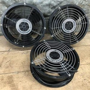 May include: Three black industrial fans with silver wire guards. Each fan has a central hub with a circular label. The fans are arranged on a wooden surface.