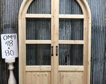 Może przedstawiać: Para drewnianych drzwi łukowych ze szklanymi panelami i czarnymi okuciami. Drzwi są wykonane z jasnego drewna i mają rustykalny wygląd. Drzwi mają 122 cm szerokości i 203 cm wysokości.