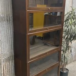 May include: A tall, dark brown wooden bookcase with five glass-fronted compartments. Each compartment has a sliding glass door and a wooden frame. The bookcase is standing next to a potted plant.
