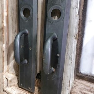 May include: Two black metal door handles with a rectangular shape and a round keyhole. The handles have a simple design and are attached to a wooden door.