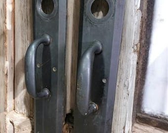 Könnte beinhalten: Zwei schwarze Metall-Türgriffe mit rechteckiger Form und einem runden Schlüsselloch. Die Griffe haben ein schlichtes Design und sind an einer Holztür befestigt.
