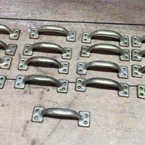 May include: A collection of 15 silver-toned metal drawer pulls with a simple, curved design. The pulls are arranged in rows on a wooden surface.
