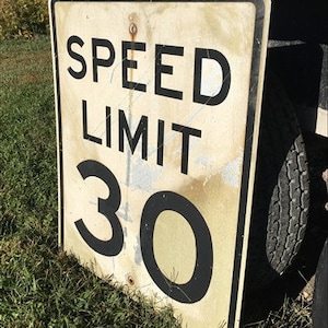 May include: A worn speed limit sign displaying "SPEED LIMIT 30" in black text. The sign has a rectangular shape with rounded corners and a black border. It is positioned on a grassy area.