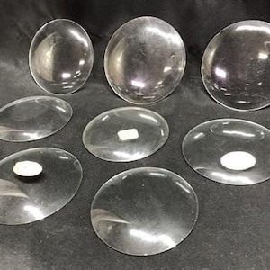 Può includere: Una collezione di lenti in vetro trasparenti, ovali e circolari, di varie dimensioni. Le lenti sono trasparenti, con una leggera curvatura, e sembrano essere realizzate in vetro lucido. La disposizione suggerisce componenti ottici o elementi decorativi.