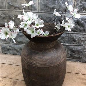 以下が含まれることがあります： 広くて浅い上部を持つ濃い茶色の木製花瓶で、白とピンクの花が入っています。 花瓶は質感のある、古びた外観をしています。 花はピンクの縁取りのある繊細な白い花びらを持っています。