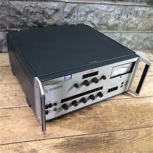 Puede incluir: Un generador de señales Cushman CE-20A-50 con una parte superior negra y una cara plateada. Cuenta con múltiples perillas, diales y un medidor. La unidad tiene un asa lateral y está sobre una superficie de madera.