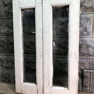 Op de afbeelding: Een paar witte geschilderde houten deuren met glaspanelen. De deuren hebben een eenvoudig ontwerp met een enkel paneel glas in elke deur. De deuren zijn in goede staat met enkele tekenen van slijtage.