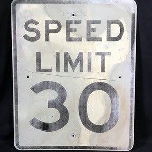 May include: A white metal speed limit sign with black text and numbers. The sign reads "SPEED LIMIT 30".