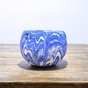 Könnte beinhalten: Ein runder, blau-weißer Marmor-Pflanztopf. Der Pflanztopf hat ein wellenförmiges Muster und steht auf einer Holzoberfläche. Der Pflanztopf ist ein dekorativer Gegenstand, der zum Halten von Pflanzen oder anderen Gegenständen verwendet werden kann.