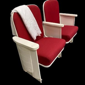 May include: Two theater seats with vibrant red fabric upholstery and off-white plastic armrests and side panels. A white lace fabric is draped over the back of the left seat. The seats feature a folding design with integrated armrests on each side.