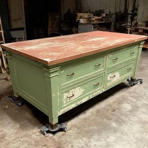 May include: A large, antique-style kitchen island with a distressed green painted finish. The countertop is a reddish-brown color. The island has multiple drawers and sits on casters for mobility. The paint is chipped, giving it a vintage look.