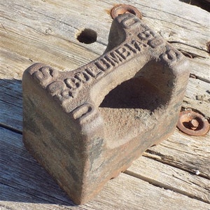 May include: A rusty cast iron weight with the inscription "50 COLUMBIA 50" on the top and sides. The weight has a handle on the top and is used for weighing items.