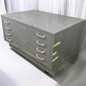May include: A large, gray metal flat file cabinet with five drawers. Each drawer has a silver handle. The cabinet is rectangular and sits on a low base with wheels. The cabinet is designed for storing large documents or artwork.