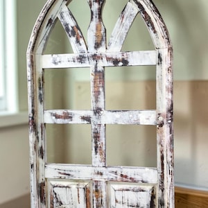 May include: A distressed, arched wooden window frame with a white finish and brown undertones. The frame features a grid pattern and decorative top, creating a rustic, vintage aesthetic. The window is likely for interior decor.