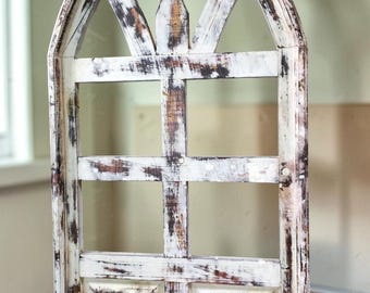 May include: A distressed, arched wooden window frame with a white finish and brown undertones. The frame features a grid pattern and decorative top, creating a rustic, vintage aesthetic. The window is likely for interior decor.