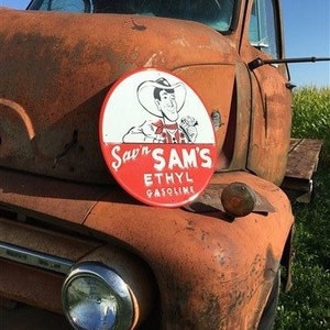 Peut inclure: Un camion vintage rouillé orange avec une enseigne ronde rouge, blanche et noire qui dit "Saw 'n Sam's Ethyl Gasoline". L'enseigne représente un cow-boy de dessin animé tenant une pompe à essence.