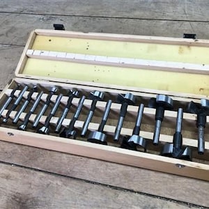 May include: A wooden box containing a set of various-sized Forstner drill bits. The bits are arranged in a light-colored wooden box with a yellow interior. The bits are made of metal and have a cylindrical shape with a flat cutting edge.