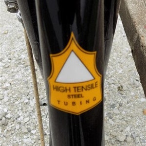 Op de afbeelding: Close-up van een zwart fietsframe met een geel en wit embleem. Het embleem leest "HIGH TENSILE STEEL TUBING" in zwarte tekst. Het frame is glanzend en reflecteert de omgeving.