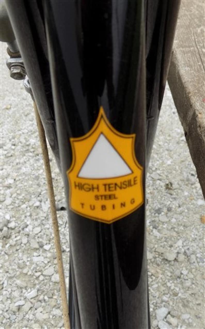 Op de afbeelding: Close-up van een zwart fietsframe met een geel en wit embleem. Het embleem leest "HIGH TENSILE STEEL TUBING" in zwarte tekst. Het frame is glanzend en reflecteert de omgeving.
