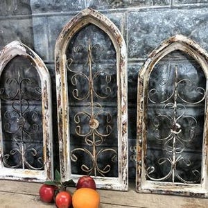 May include: Three arched window frames with distressed white and brown paint finishes. Each frame contains a central vertical metal rod with symmetrical, decorative scrollwork patterns. The frames are positioned upright in a row.