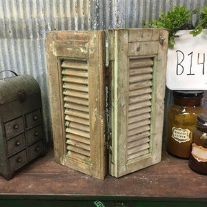 May include: A pair of weathered wooden shutters with louvered panels, painted in a light brown color. The shutters are hinged together and have a rustic, vintage look. The shutters are displayed on a table with a metal box and glass jars.