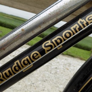 Op de afbeelding: Detail van een vintage fietsframe. Het zwarte frame heeft gouden letters met de tekst "Rudge Sports". Een zilveren metalen buis is diagonaal over het frame geplaatst. De achtergrond toont een wazig groen veld en lucht.