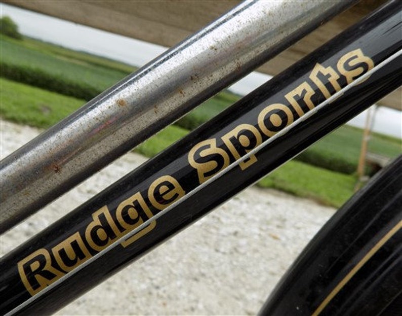 Op de afbeelding: Detail van een vintage fietsframe. Het zwarte frame heeft gouden letters met de tekst "Rudge Sports". Een zilveren metalen buis is diagonaal over het frame geplaatst. De achtergrond toont een wazig groen veld en lucht.