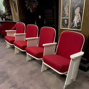 May include: A row of vintage theater seats with red fabric cushions and white armrests and frames. The seats are arranged in a row, suggesting a classic cinema or auditorium setting. The seats have a retro design.