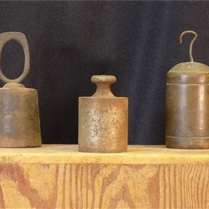 May include: Three vintage metal weights, one with a ring handle, one with a hook, and one with a smooth top. The weights are sitting on a wooden shelf.