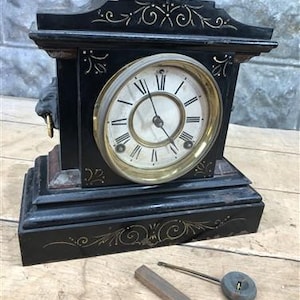 May include: A black mantel clock with gold-toned floral detailing and a circular face featuring black Roman numerals. The clock has a brass bezel, side lion head ornaments with rings, and is accompanied by a separate metal pendulum and a wooden bar.