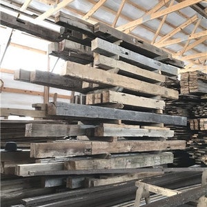 Puede incluir: Una pila de vigas de madera recuperada, algunas con nudos e imperfecciones, en un almac&eacute;n. Las vigas son de varios tonos de marr&oacute;n, gris y blanco, y est&aacute;n apiladas de forma desordenada.