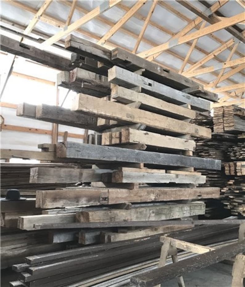 Puede incluir: Una pila de vigas de madera recuperada, algunas con nudos e imperfecciones, en un almac&eacute;n. Las vigas son de varios tonos de marr&oacute;n, gris y blanco, y est&aacute;n apiladas de forma desordenada.