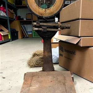 May include: A vintage Toledo industrial floor scale with a large circular dial face featuring the text TOLEDO NO SPRINGS HONEST WEIGHT. The scale has a weathered brown metal finish, a rectangular platform base, and a vertical column support.