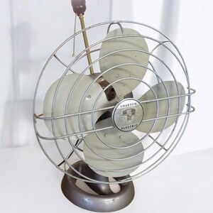 Puede incluir: Un ventilador vintage plateado Emerson Electric con una bombilla y una cadena de tracci&oacute;n. El ventilador tiene tres aspas y una jaula de metal.