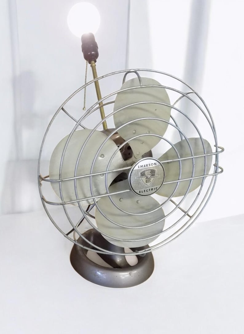 Puede incluir: Un ventilador vintage plateado Emerson Electric con una bombilla y una cadena de tracci&oacute;n. El ventilador tiene tres aspas y una jaula de metal.
