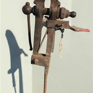 May include: A vintage cast iron post vise featuring a dark, weathered, and rusted metal finish. The tool includes a long vertical mounting leg, a horizontal screw mechanism with a rounded handle, and a pair of heavy, textured gripping jaws.