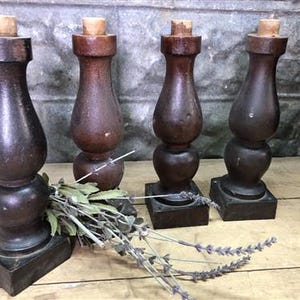 May include: Four antique wooden bottle stoppers with a dark brown finish. Each stopper has a square base, a turned body, and a cork top. A sprig of dried lavender rests in front of the stoppers.