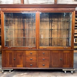 May include: A large, antique wooden display cabinet with glass doors and shelves. The cabinet has a dark brown finish, drawers, and doors. It sits on small wheels. The top of the cabinet has decorative molding.