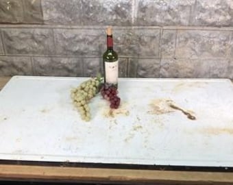 Könnte beinhalten: Ein weißes Schneidebrett mit schwarzem Rand, auf dem sich eine Weinflasche und Trauben befinden. Das Brett hat einen abgenutzten, rustikalen Look mit sichtbaren Flecken.