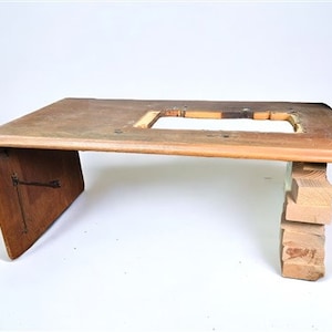 May include: A wooden table base with a rectangular top and a cutout in the center. The base is made of several pieces of wood that are stacked on top of each other. The table is unfinished and has a natural wood color.
