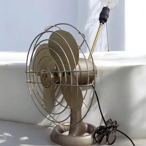Puede incluir: Un ventilador vintage de metal plateado con un cable negro y una bombilla en la parte superior. El ventilador tiene una jaula de metal circular y tres aspas. El ventilador est&aacute; etiquetado como "Emerson Electric".