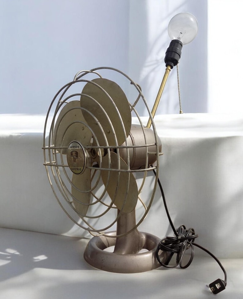 Puede incluir: Un ventilador vintage de metal plateado con un cable negro y una bombilla en la parte superior. El ventilador tiene una jaula de metal circular y tres aspas. El ventilador est&aacute; etiquetado como "Emerson Electric".