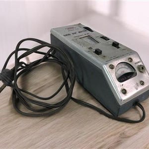Op de afbeelding: Een vintage zilver- en grijze Grid Dip Meter met een zwart netsnoer en stekker. Het apparaat heeft een wijzerplaat, knoppen en een frequentiebereikindicator. De meter staat op een lichtgekleurd houten oppervlak.