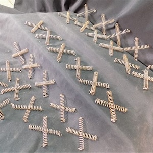 May include: A collection of silver metal spring clips, arranged in a pattern on a gray fabric surface. The clips are shaped like a cross and are made of coiled wire.
