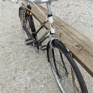 Op de afbeelding: Een vintage zwarte en zilveren fiets met witte stuur en een zwart zadel. De fiets heeft een klassiek ontwerp met een gebogen stuur en een voorspatbord. De fiets staat op een grindoppervlak.