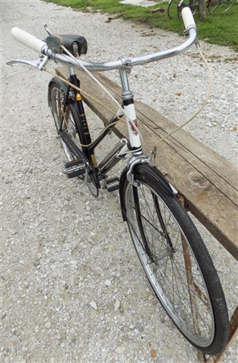 Op de afbeelding: Een vintage zwarte en zilveren fiets met witte stuur en een zwart zadel. De fiets heeft een klassiek ontwerp met een gebogen stuur en een voorspatbord. De fiets staat op een grindoppervlak.