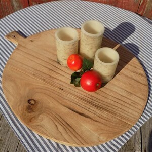 May include: A round wooden serving board with a handle, featuring a natural wood grain pattern. Three white candles and two red apples are arranged on the board.