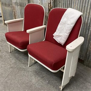 May include: Two vintage theater chairs with red fabric seats and white armrests and frames. One chair has a white, patterned cloth draped over the back. The chairs are set against a corrugated metal wall.